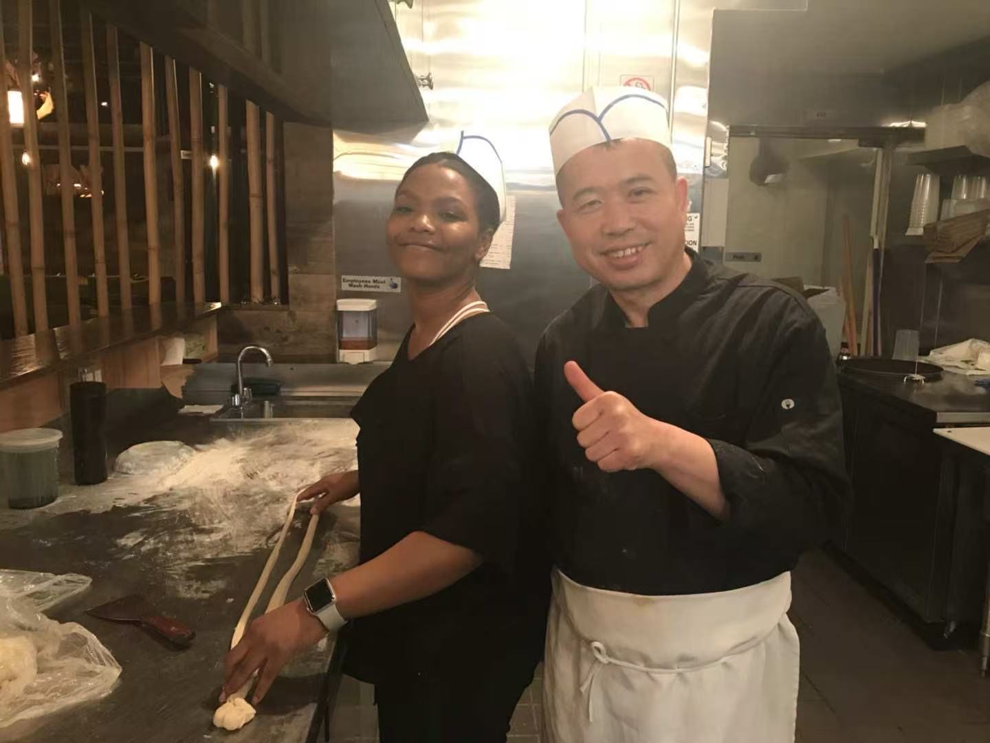 Apprentice rolling noodle strands with master smiling beside
